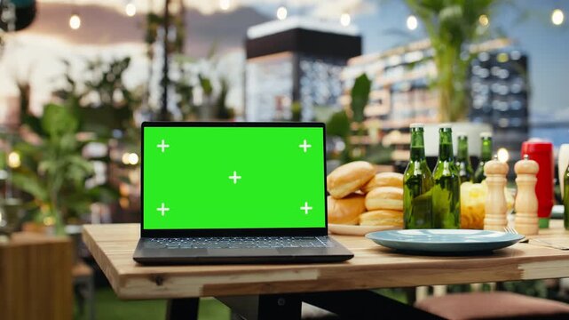 Chroma key laptop mockup sits on a rooftop party dining table with food and drinks, providing an editable display for screen replacement at an outdoor event.