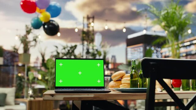 Laptop displaying a green chroma key screen stands beside beer bottles, bread and plates on a rooftop barbecue table, offering an editable computer display for an outdoor grill party.