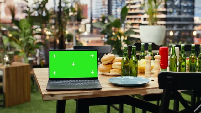 Laptop with a green chroma key screen sits beside beer bottles, bread and plates on a rooftop barbecue table, providing an editable display for screen replacement at an outdoor grill party.