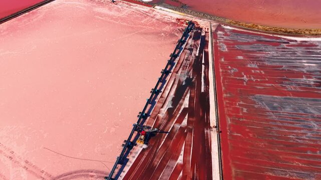 Heavy harvester machine extracting resources from pink salt flat. Top down perspective highlights stark contrast between bright crystallized bed and dark harvested lines.