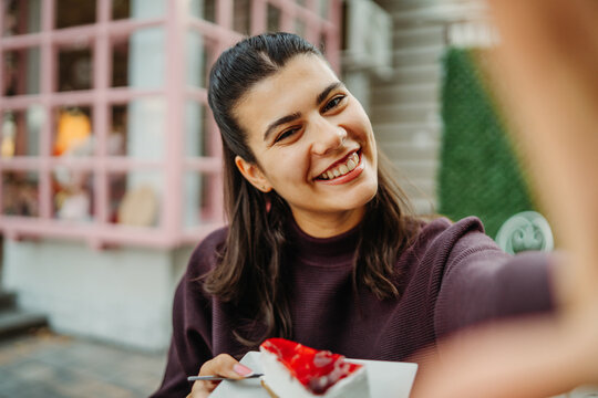 Smiling woman taking a selfie while holding cake at an outdoor cafe