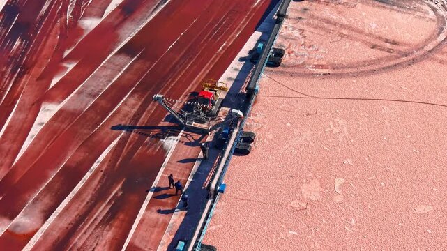 Tracked extraction equipment operating on bright pink salt bed. Aerial camera captures industrial harvesting process leaving distinct parallel brown trails.