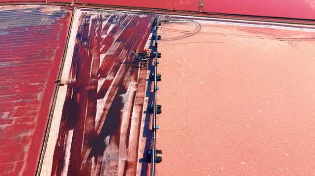 Wide aerial shot of continuous salt mining machinery in red lake. Long conveyor system systematically strips mineral layer exposing dark earth underneath.