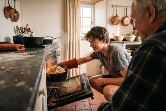 Smiling teenage boy taking freshly baked sourdough bread out of the hot oven while his grandfather watches in a cozy rustic kitchen