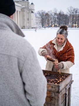 Smiling mature woman street vendor scooping hot roasted chestnuts into a paper cone for a customer in a snowy European city square during winter