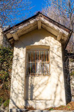 View of a small, weathered roadside shrine featuring a statue behind a rusted metal grate, set against a stone wall with bare tree branches, Montvernier, Auvergne-Rhone-Alpes, France.