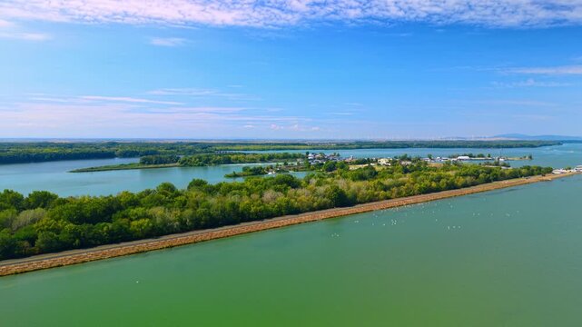 Long straight river embankment separating green water. Drone perspective highlights linear stone and grass dike with paved road dividing large reservoir under clear sky.