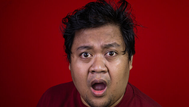 A comical close up portrait showing a young Asian male with messy dark hair displaying a highly exaggerated shocked expression. The amazed adult stares directly forward with wide open