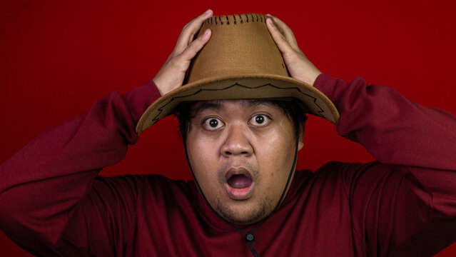 An astonished young Asian male holding the top of his brown cowboy hat with both hands while staring forward with wide eyes and a dropped jaw. The incredibly shocked subject is dressed