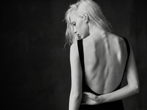 Black and white artistic portrait of blonde woman in backless black top looking down against dark background