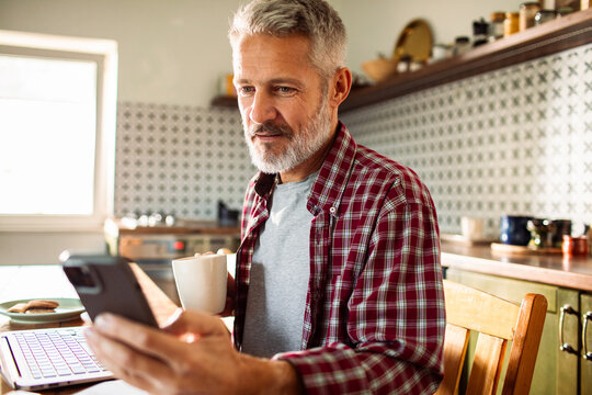 Middle-aged man checking smartphone with coffee in kitchen