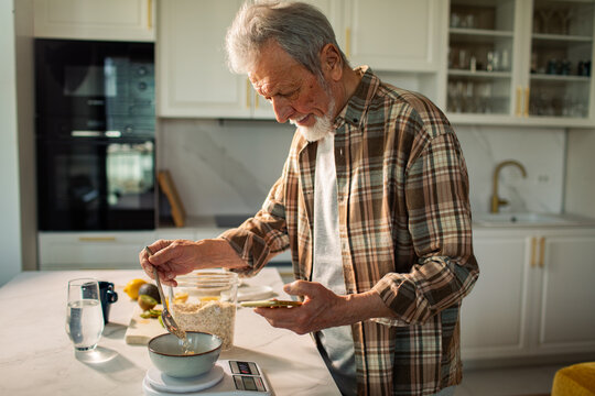 Senior man weighing oatmeal on kitchen scale while using smartphone