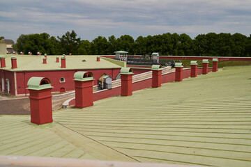 Historic fort rooftop scene with green metal roofing panels, red brick chimneys, defensive parapet walkway, and cannon fixtures beneath cloudy sky © diczman