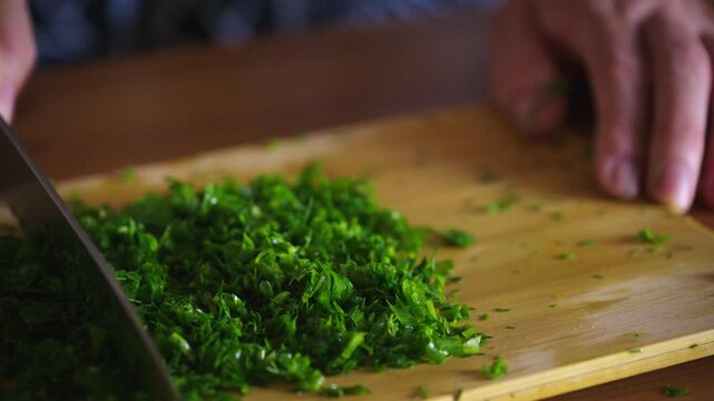 Fresh parsley chopped finely on wooden board. Hands guide knife with practiced precision. Crisp greens scattered across light wood surface. Prep work for dishes, garnishes, or seasoning
