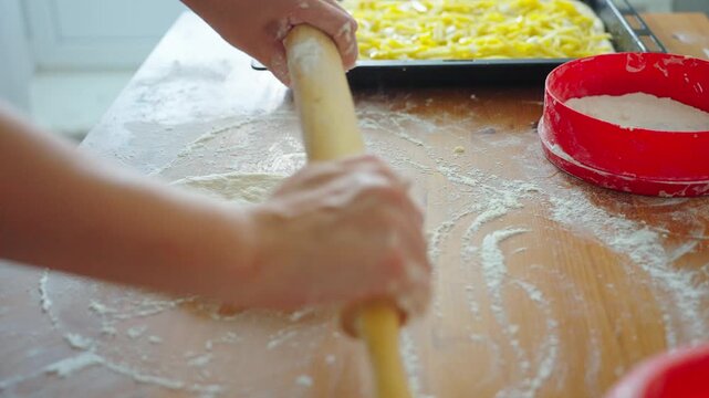 Hands roll dough on a floured wooden board. Child works calmly in a kitchen. Pizza ingredients nearby on the counter. Ideal for cooking or home kitchen scenes