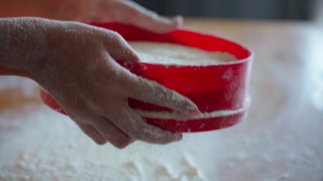 Hands sift flour into a red sieve. Dust settles softly on wooden surface. Warm, cozy kitchen scene. Perfect for baking tutorials or food prep videos