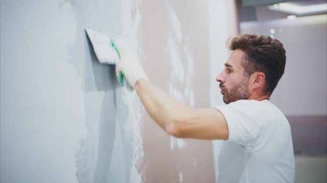 Man applies fresh plaster to a wall surface. He wears white gloves, focused on his task. The wall shows patches of old paint. His expression is concentrated, steady