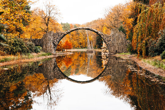 View of The Devil's Bridge in Gablenz surrounded by vibrant autumn foliage reflecting in the calm water of the Rakotzsee, Gablenz, Sachsen, Germany.