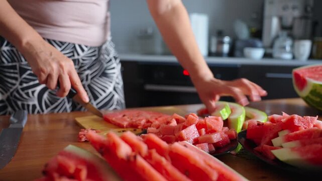 Woman slices watermelon on wooden board. Fingers guide knife with care and precision. Red fruit pieces pile up on cutting board. Kitchen background shows modern appliances