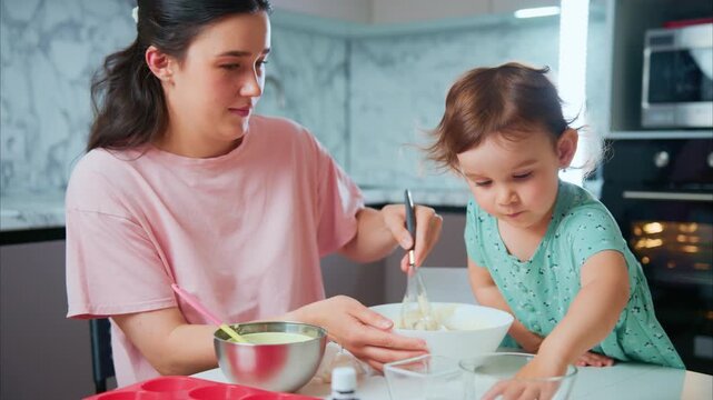 Mother smiles while guiding toddler's mixing bowl. Child focuses intently on whisking batter with tiny hands. Kitchen backdrop shows appliances and light streaming in
