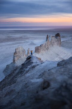 View of a person standing with arms outstretched on the rocky ridge of Dragons Crest in Bozhira, overlooking the vast desert landscape at sunset, Karakiya District, Mangystau, Kazakhstan.