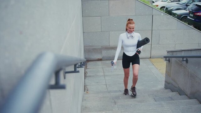 Young woman smiles as she walks down concrete stairs. She carries a rolled yoga mat and headphones. Urban setting with parked cars and stone walls. Perfect for fitness, wellness