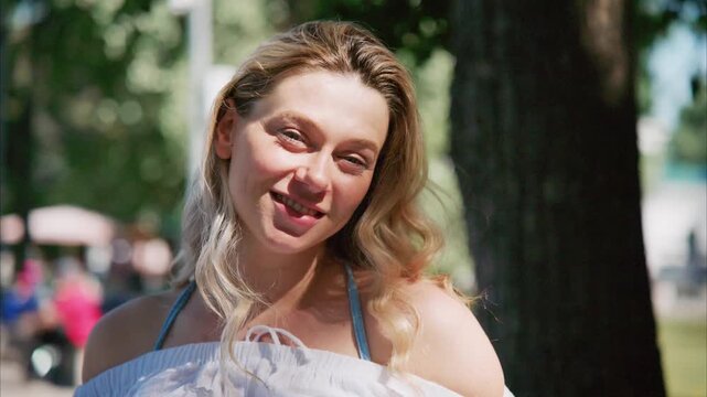 Golden hair sways in summer breeze. Woman smiles softly, eyes closed. She touches her hair, relaxed and serene. Sun-dappled park, people in background. Perfect for lifestyle, fashion, or travel scenes
