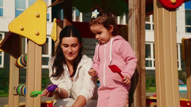 Warm sunlight glows on a wooden play structure. Woman and child share playful moments together. They smile while handling colorful toys nearby. Children enjoy outdoor playtime in a safe, bright space
