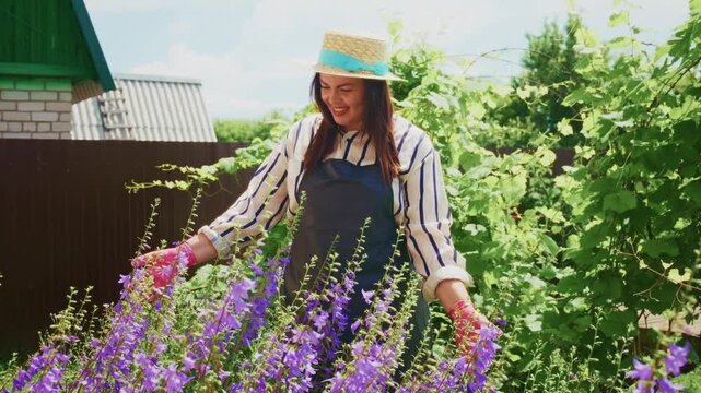 Woman smiles while tending vibrant purple flowers. She wears a hat and gardening gloves. Green vines and wooden fence frame the scene. Bright sunlight highlights her joyful gardening