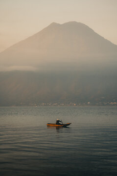 View of a lone fisherman in a wooden boat on Lake Atitlan with a volcano rising through morning mist in the background, Solola Department, Guatemala.