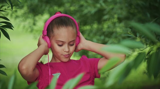 Little girl enjoys music outdoors. She wears bright pink headphones. Her pigtails sway gently as she listens. Green leaves frame her peaceful expression. Soft sunlight enhances the natural background