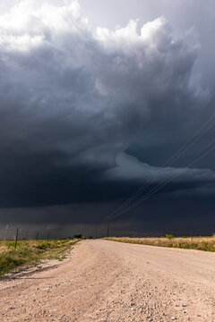 View of a dirt road leading towards a dark, ominous storm cloud with heavy rain and power lines in the world.