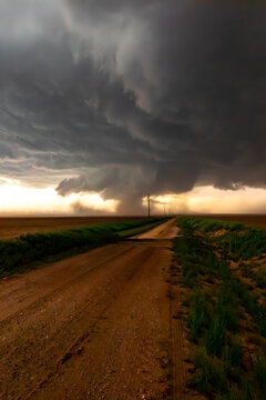 View of a powerful tornado funnel descending from dark, ominous storm clouds over a flat, rural landscape with a dirt road stretching into the distance.
