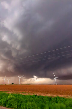 View of wind turbines standing in a field under a dark, dramatic storm sky with heavy clouds and strong winds, World.