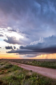 View of a dramatic storm with dark clouds and heavy rain over a vast, flat landscape with a dirt road at sunset.