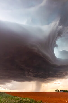 View of a dramatic supercell storm with a funnel cloud hanging over a vast red soil field under a dark, swirling sky.