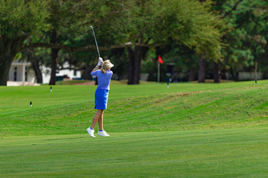 Female Golfer Swinging Iron Club on Green Golf Course Fairway Toward Putting Green Flag