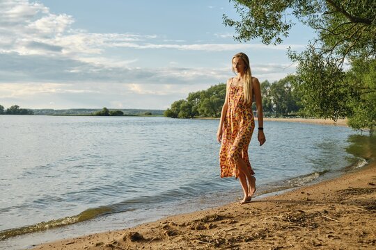 A woman in a colourful sundress strolls barefoot along the sandy shore of a tranquil lake. Lush green trees frame the water under a bright blue sky with fluffy clouds