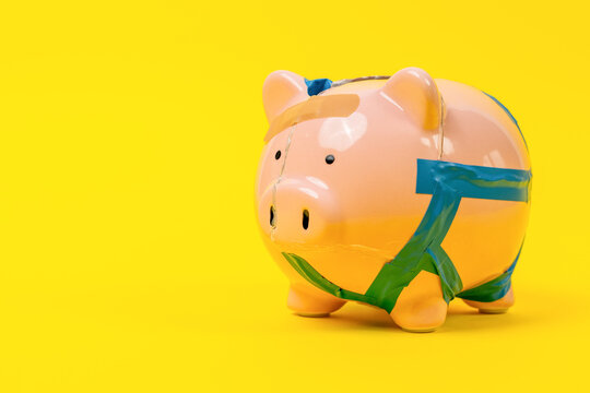 A cracked pink ceramic piggy bank mended with blue and green adhesive tape and a sticking plaster stands on a bright yellow studio backdrop, a concept of financial crisis and recovery with space for.