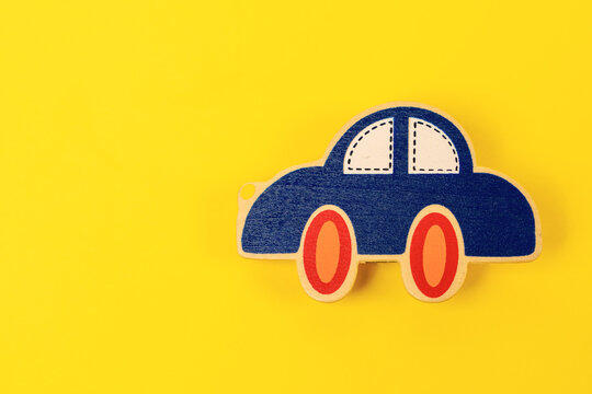 A small blue wooden toy car with red and orange wheels and white windows rests on a bright yellow surface with empty space on the left, a concept of childhood play, mobility and auto insurance.