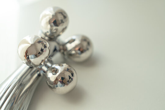 Four faceted stainless steel balls on a handheld cosmetology gadget rest near a bright window on a pale desktop, a minimalist idea of modern home beauty technology and daily grooming ritual.