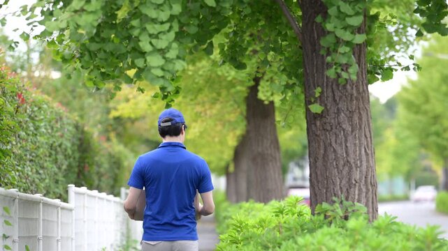 新緑の屋外で段ボール箱を持って荷物を運ぶ歩く配達する若い日本人男性の動画　物流や配送業で働く配達スタッフイメージ　顔無しの後姿