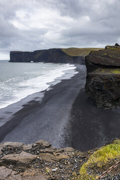 View of the dramatic black sand beach and towering basalt cliffs along the South Coast under a cloudy sky in Dyrholaey, Myrdalshreppur, Iceland.