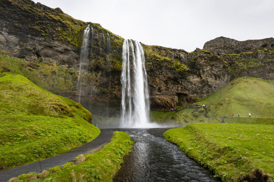 View of Seljalandsfoss waterfall cascading over rocky cliffs into a stream with lush green grass and people walking along the path in Seljalandsfoss, Rangarthing eystra, Iceland.