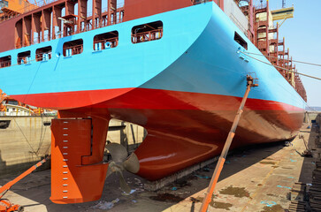 Rear view of a merchant ship in dry dock after fresh hull painting, showing the stern, rudder and large propeller exposed above the dock floor during shipyard maintenance and inspection. © Mariusz