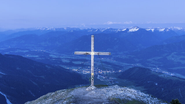 View of the summit cross on Stoderzinken overlooking the Ennstal valley near Schladming at twilight with distant mountain peaks and valley lights, Stoderzinken, Steiermark, Austria.