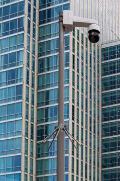 View of a security camera mounted on a metal pole in front of a modern glass skyscraper in Almere, Flevoland, Netherlands.