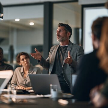 Bearded man in a grey blazer gestures while presenting to colleagues at a glass-walled conference table with laptops.