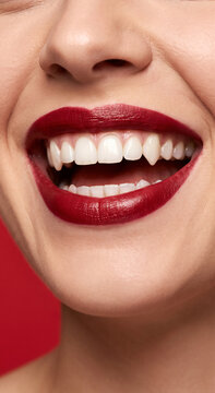 Close up of woman smiling with bright red lipstick and vampire fangs, dark red background. Halloween costume, horror, makeup, fantasy.