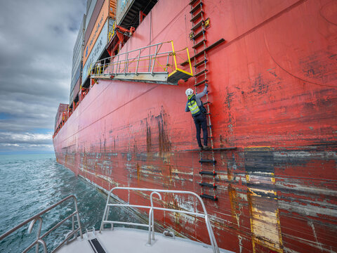 View of a pilot climbing a ladder on the side of a large red container ship during shipping operations at Port of Tauranga, Tauranga, Bay of Plenty Region, New Zealand.